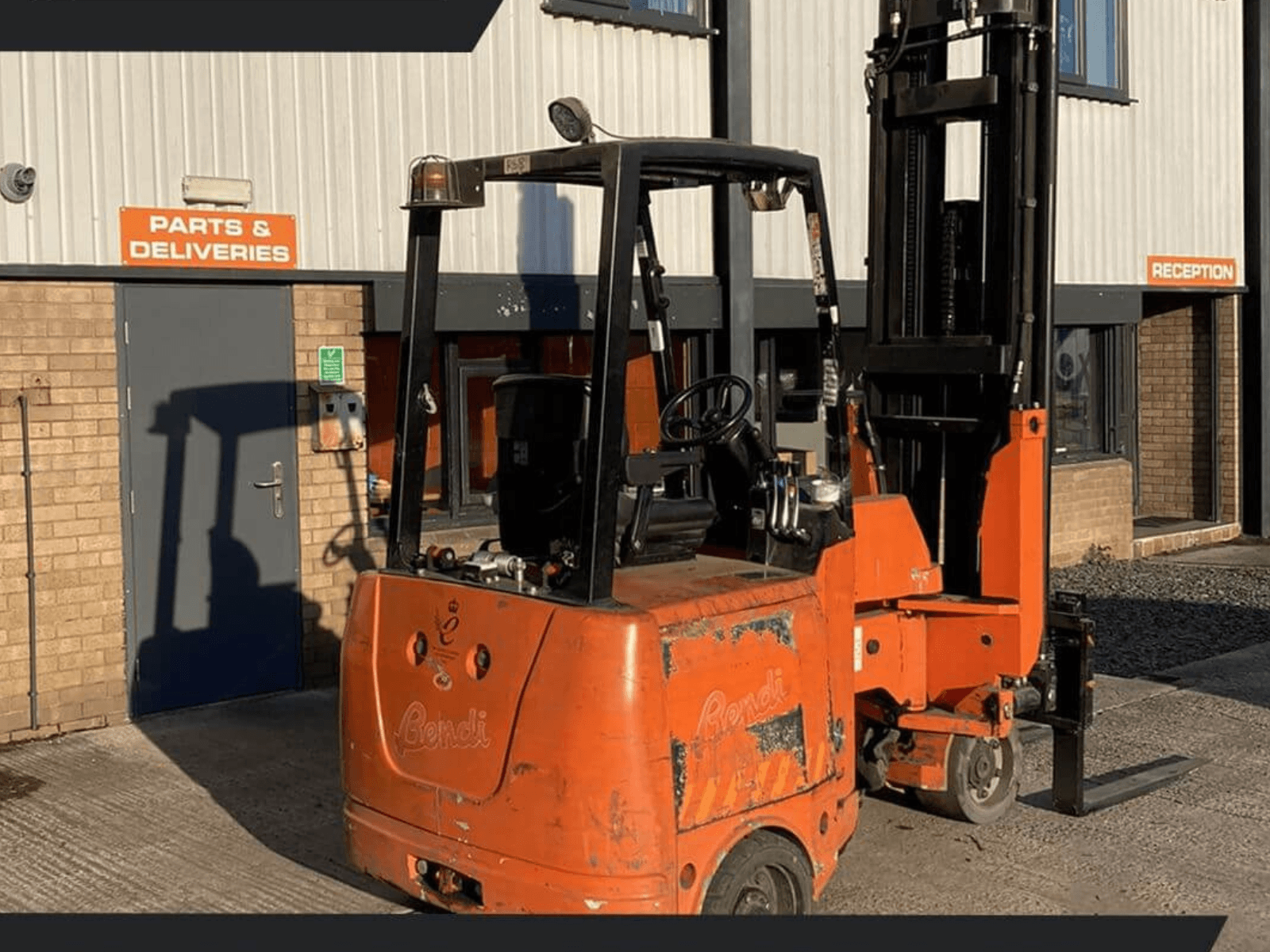 A bright orange industrial forklift positioned outside a warehouse with parts and deliveries signage.