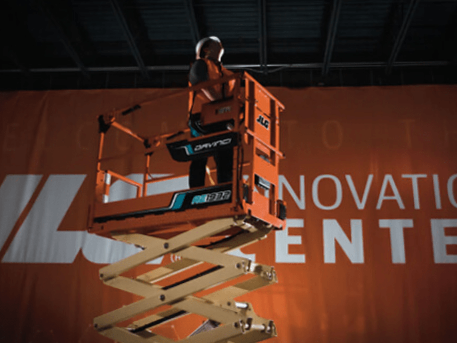 A worker in safety gear on a scissor lift, positioned in front of a large orange banner that reads 'INNOVATION CENTER'.