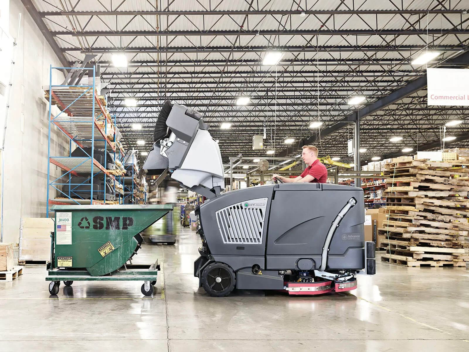 A person operating an industrial floor scrubber next to a green waste container in a warehouse setting with shelves and pallets in the background.