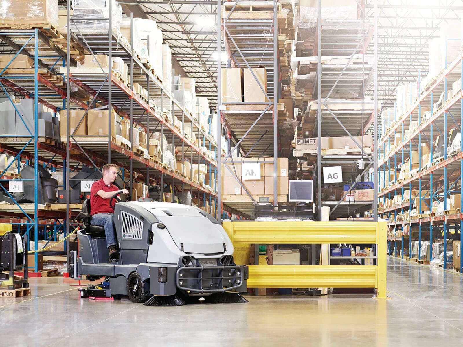 A warehouse environment featuring a worker operating a floor scrubber amidst shelves of stored products.