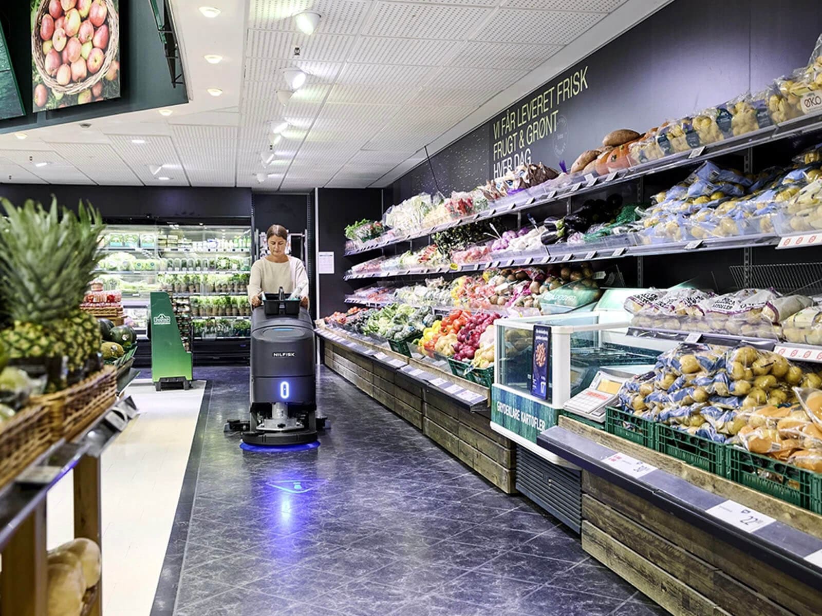Interior of a grocery store showcasing fresh produce with an employee using a floor cleaning robot.
