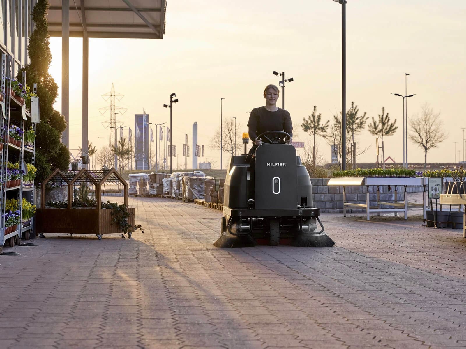 A person operates a cleaning machine on a well-paved outdoor area of a store during sunset.